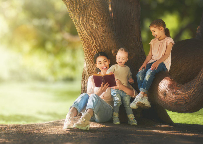 Mother and daughters sitting under the tree on summer lawn. Happy family playing outdoors. Pretty young mom reading a book to her children in the park outside